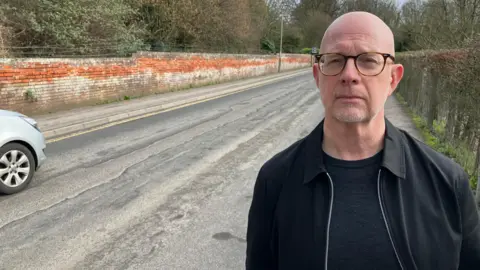 A bald man with glasses wearing a black t-shirt and black unzipped jacket stands next to station road in Devizes where there is large scale visible degradation to the road surface beside him