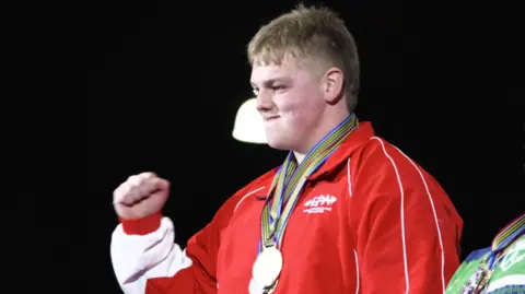 Will Anders, who has medium length, light brown hair. He is wearing a red tracksuit and wearing medals. He is biting his lip and fist pumping the air in celebration.