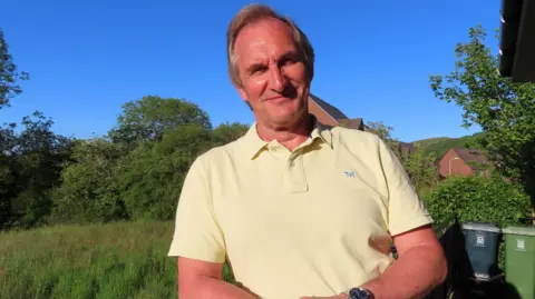 Mike Mann Mike Mann has short grey hair and is wearing a yellow polo shirt. He is leaning on a fence in a garden while smiling.