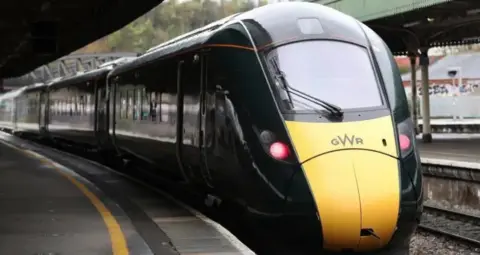 PA Media Yellow front of train coming towards the camera with two red lights lit and dark green carriages behind -railway platforms either side.