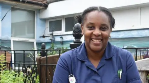 A nurse with short black hair and wearing a blue shirt sits on a wooden bench i front of a blue and white hospital building with two small windows to the left. She has an ID badge pinned to her shirt to the left and two pens in her shirt pocket to the right. There is a black railed gate between the building and the bench.