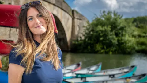 A woman with long light brown hair, with sunglasses on her head, wearing a dark blue T-shirt. She is smiling, stood in front of a river with some canoes moored up. There's a bridge and some trees in the background. 