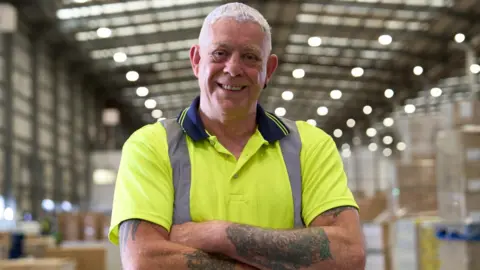 Ken Wheatley - a man with short white hair and wearing a bright yellow t-shirt - smiles as he stands in a large warehouse with his arms folded.