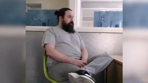 A man sitting in a green chair, photographed from the side. He has long brown hair in a bun and a long brown beard and moustache. He is wearing a grey T-shirt and grey tracksuit bottoms. He is looking off to the right, talking to someone across a police desk. The photo was taken during Caradain's police interview.