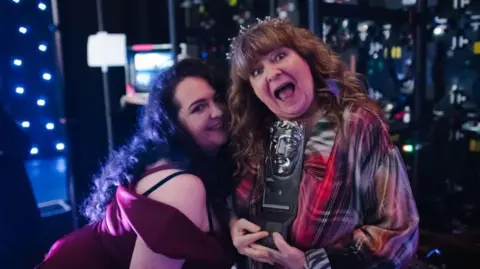 Getty Images Janey Godley with daughter Ashley Storrie in November 2019. Ashley stands side on leaning into her mother's shoulder, smiling - she wears a red velvet dress and has long dark curly hair. Godley is dressed in plaid with long curly brown hair and is holding a Bafta award - she has her mouth open in celebration