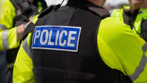 Close up of the back of police officer's black jacket, which has the letters police in white written on. There are two other officers in the distance but you cannot see their faces.