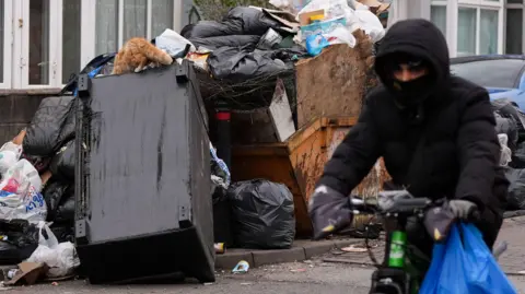 A cyclist passes an overflowing pile of rubbish. A cat is on top of the rubbish looking through it. The pile includes an overturned sofa and a skip, overflowing with rubbish. The cyclist wears a black coat and has a black scarf over his mouth while he has a blue plastic bag in one hand as he cycles.