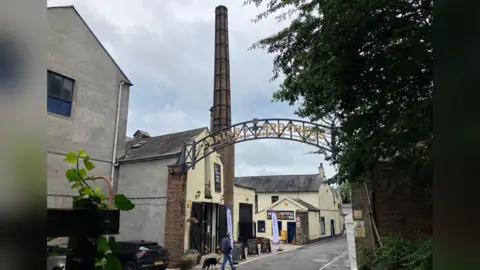 The exterior of Jennings Brewery. A tall brick chimney rises out of a two-storey yellow building, which has multiple signs on it. A metal arch over the entryway reads: "Jennings Brothers".