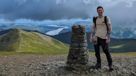 Supplied Mr Kendall is pictured on top of a mountain in the Lake District. He stands next to a cairn