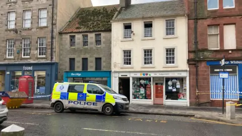 A blue, yellow and white Police Scotland van parked outside a shop with a flat upstairs where the windows are open. There is police tape at the right and left hand sides a red phone box on the left hand side.