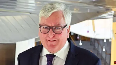 A man with short white hair and glasses smiles to the camera. He is in the Scottish Parliament foyer and wears a dark suit, white shirt and black tie. 