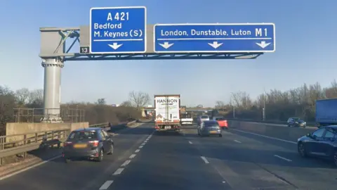 The exit for junction 13 of the M1 southbound, showing four lanes of traffic and a big sign above the road showing the right three lanes for the M1 - and the inside lane for the A421.