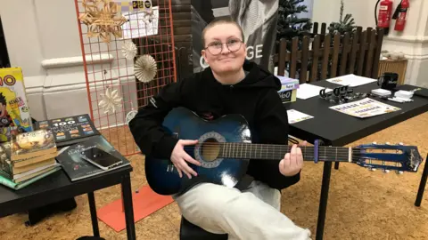 Olly Armstrong posing with a guitar in 2 Royal Avenue, Belfast.  He has shaved hair and glasses and is wearing a black hooded sweatshirt and light-coloured trousers. 