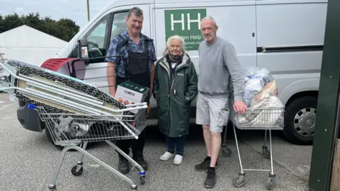 Two middle aged men and an older woman stand in front of a large silver van marked Hidden Help 