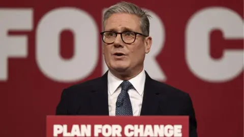 PA Media Sir Keir Starmer, with grey hair and wearing dark glasses, speaks at a podium in front of a red background 