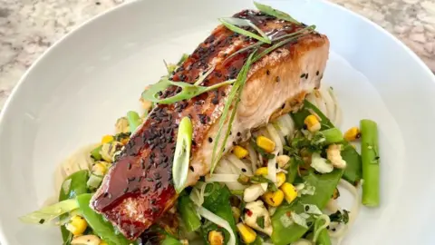Handout A piece of fish is resting on top of a bed of green vegetables, sweetcorn and noddles. 