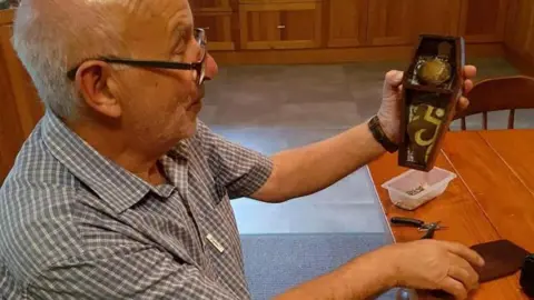 Family photograph Image of an older man siting at a table, holding a small coffin-shaped box in his hands.