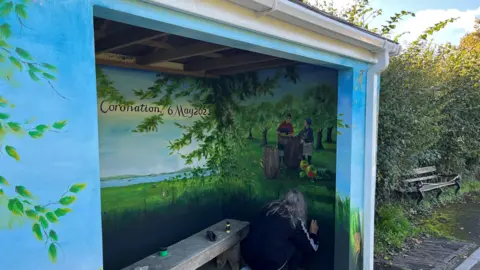 We are looking at a bus shelter which has been transformed into a beautifully decorated scene of the countryside. In the corner a grey-haired woman is adding some touches of paint to the inside wall of the bus shelter