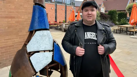 Gavin Kermack / BBC A young man wearing a black cap, a black t-shirt and a black leather jacket standing next to the above artwork.