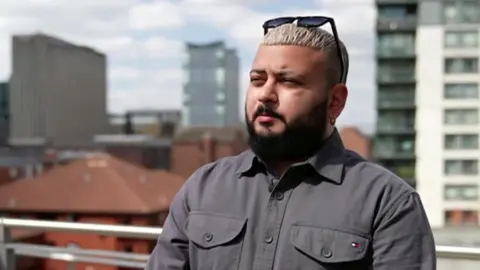 Ankush Dupar stands outdoors on a city-centre rooftop terrace. He is wearing a dark grey buttoned shirt and sunglasses rest on top of his head. Office and apartment buildings can be seen in the background. 