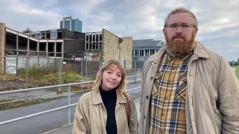 A shorter, blond-haired woman wearing a beige mackintosh standing next to a taller, bearded man with ginger hair and wearing a beige coat with a yellow, blue and white checked shirt. Derelict school buildings loom behind metal fences in the background.  