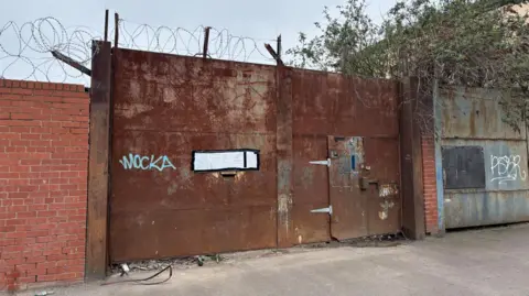 Simon Thake/BBC A large brown metal gate with barbed wire above it. A white notice is taped to the gate.