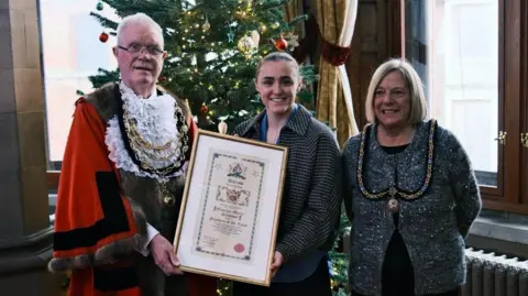 Ian Allington Mayor of Barrow Frederick Chatfield, who has short white hair, wears red robes and mayoral chains, presents Georgia Stanway with her Freedom of the Town framed certificate. It looks very formal with traditional ink writing. She is smartly dressed in a blue blouse, chequered jacket and has brown hair tied back. Mayoress Elizabeth Chatfield is on the other side of her. She has shoulder length blonde hair and wears a glittery grey cardigan, black top and a mayoral chain. There is a Christmas tree behind them. All three are smiling.