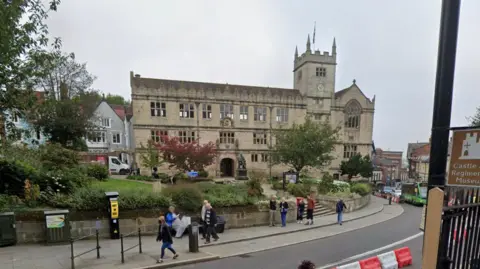 An old building with a statue of Charles Darwin sat in front of it. The building it sat back from the road, with people walking past on the path