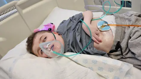 Handout/PA Real Life A young boy in a hospital bed wearing an oxygen mask