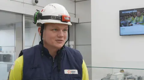 David Upex is wearing a yellow top, blue, EDF gilet and a white, hard hat with a torch and ear defenders attached. There is a TV screen on a wall behind him.