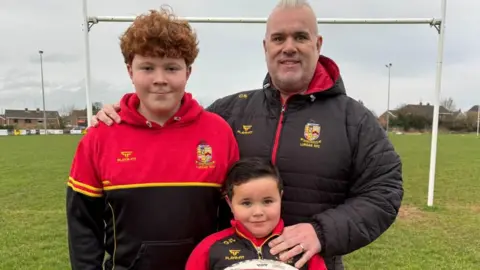 Ollie smiles holding a red and white rugby ball at Lurgan rugby pitches. His dad Colin has his hand on Ollie's shoulder and is wearing a black coat. Colin also has his arm around his son Niall who has red hair and is smiling. Niall is wearing a red and black hoody.