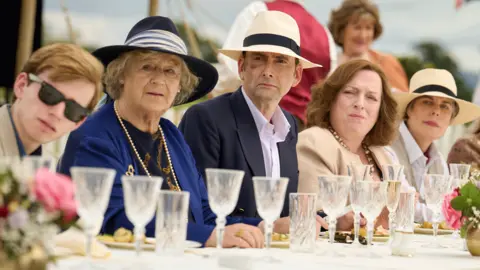 Disney+ A scene from Rivals as guests watch a game of polo over lunch. They sit at a table lined with champagne flutes, and flowers, and the men and women wear straw hats and blazers and formal jackets. 