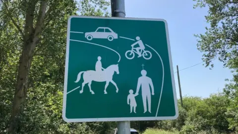 A traditional quiet lanes sign. It is green with images of a pedestrian, horse rider, cyclist and car.