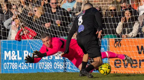 Lyn & Trev Sports A goalkeeper dives to try and stop a ball from being kicked into a net. A packed crowd can be seen in the background, behind a white net. 