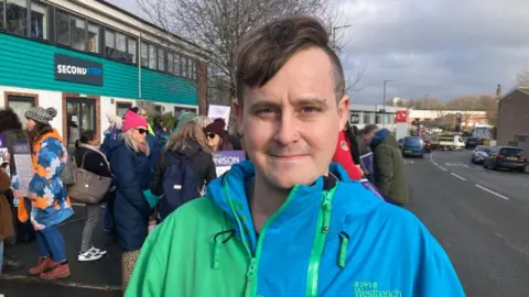 Daniel King, a man with dark hair, piercings and a blue and green raincoat, stands on a Unison picket line outside Second Step HQ in Bristol.