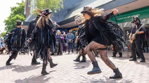 Neil King A group of morris dancers wearing all black with top hats and dancing around