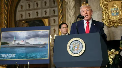 Donald Trump speaking at a podium in Mar-a-Lago. He is wearing a dark navy blue suit with a bright red tie. Behind him is Secretary of State Marco Rubio. Beside him is a big photo featuring a rendering of Trump Class USS Defiant. Hie is standing in a room with ornate, gold accents behind him. 