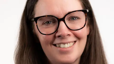 Katie Warburton A professional headshot of Katie. She has long dark hair, blue and green eyes and is wearing dark rimmed glasses. She is smiling at the camera.