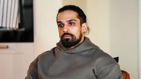 A still from EastEnders showing the character of Ravi, wearing a grey hoodie and looking into the distance.