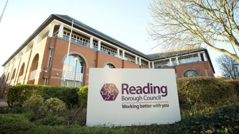 A brick building with a hedge outside, with a large white and purple sign saying 'Reading Borough Council, working better with you'.