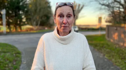 A woman in a white jumper stand by a road outside her house. 