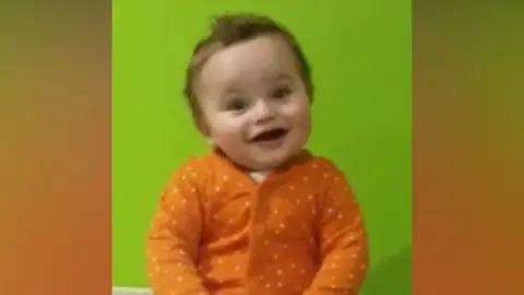 Baby in orange and white polka dot baby grow smiling while sitting on a changing table. Green wall behind him. 