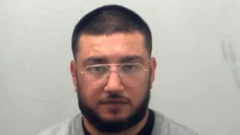 Essex Police A man in a custody photo. He has a beard and glasses and has a beard. He is looking at the camera 