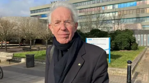 BBC A man with white hair and glasses and wearing a black coat and scarf standing in front of a grey office building 