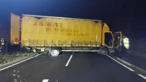 A yellow lorry is sideways and is across a road. Its back doors and open and the drivers side door is open. Two police officers are standing at either end of the vehicle.