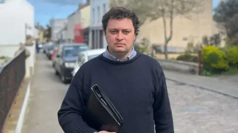 Alex Snowdon is wearing a navy knitted jumper over a light blue shirt as he stands on a cobbled street while carrying a large black folder. 