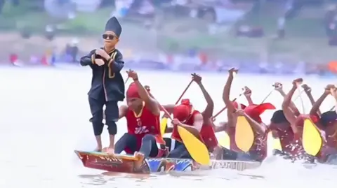 A boy, dressed in black wearing sunglasses, standing on the tip of a racing boat while a dozen adult atheles, in red uniforms, rowing behind him. 