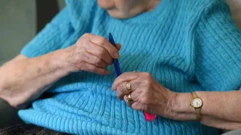 An elderly person is sitting in a chair holding a pen. The person is wearing a gold watch and gold ring fingers. She is holding a blue pen in her hands. She is wearing a blue cable knit top.