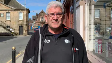 A man in a black top and jacket stands outside a shop with a historic Tolbooth building in the background