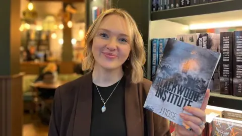 Alice Cullinane/BBC A woman with short blonde hair is wearing a black top and a brown blazer. She is holding a book that says 'Blackwood Institute'. 